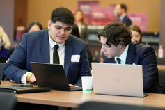 Two male graduate students look at laptops in a classroom.